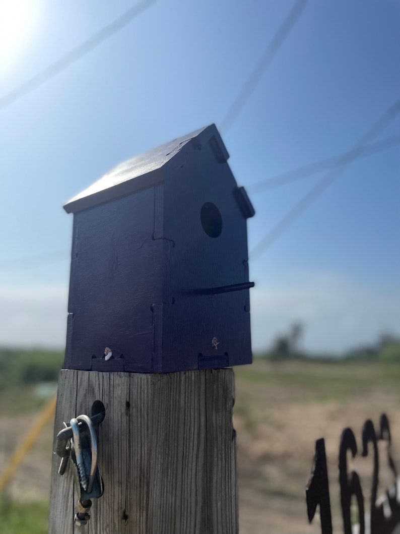 Puede incluir: Una casita de p&aacute;jaros azul oscuro se asienta sobre un poste de madera. La casita de p&aacute;jaros tiene una peque&ntilde;a abertura para que los p&aacute;jaros puedan entrar. El poste est&aacute; desgastado y tiene un gancho de metal sujeto a &eacute;l.