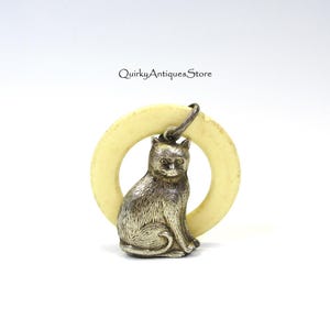 May include: A silver cat pendant with a cream-colored ring. The cat is detailed with visible fur and facial features. The ring is a pale yellow color. The text "Quirky Antiques Store" is visible in the upper right corner.
