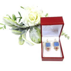 May include: A pair of gold-toned earrings with blue square stones and small red stones, presented in a red jewelry box. The earrings are displayed against a white background, with a floral arrangement in the upper left corner.
