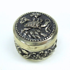 May include: A silver trinket box with an ornate floral design. The lid features a raised, detailed floral motif with a textured center.