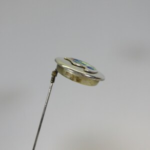 May include: A silver-toned hat pin with a circular head. The head features a colorful design with blue, green, and yellow accents. The pin is angled, showing the details of the design and the pin's construction.