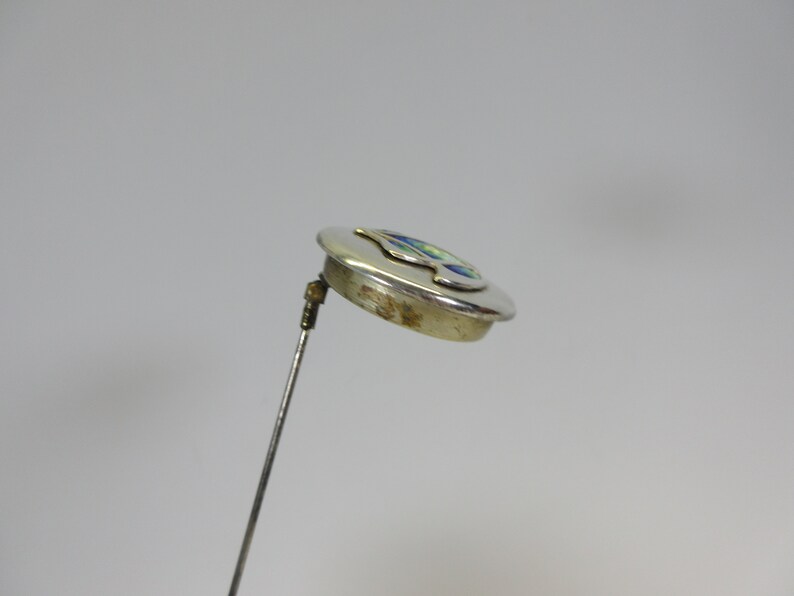 May include: A silver-toned hat pin with a circular head. The head features a colorful design with blue, green, and yellow accents. The pin is angled, showing the details of the design and the pin's construction.