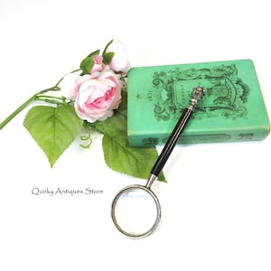 May include: A vintage magnifying glass with a black handle and silver-toned accents rests on a green book. Pink roses and green leaves are arranged nearby. The magnifying glass has a circular lens and ornate detailing.