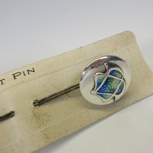 May include: A silver hat pin with a round head featuring a blue, green, and white design. The pin is on a vintage cardboard backing that reads "HAT PIN".