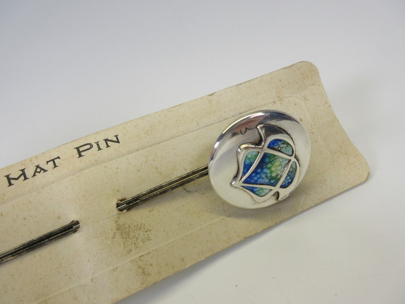 May include: A silver hat pin with a round head featuring a blue, green, and white design. The pin is on a vintage cardboard backing that reads "HAT PIN".