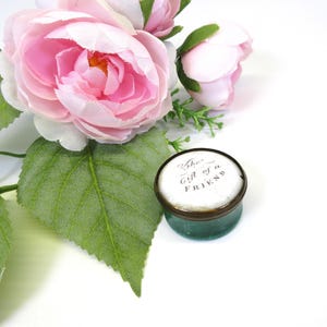 May include: A small, round, lidded container with a dark green base and a white top featuring the black printed text The Gift of a FRIEND, positioned next to a cluster of artificial light pink roses with green leaves on a white background.