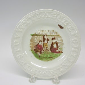 May include: White ceramic plate with a raised alphabet border. The center of the plate features a hand-painted scene of three boys playing a game with a ball. The scene is surrounded by a green border.