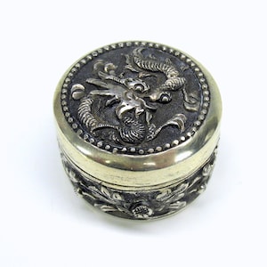 May include: A silver trinket box with an embossed design of a dragon on the lid. The box is decorated with floral patterns around the sides.