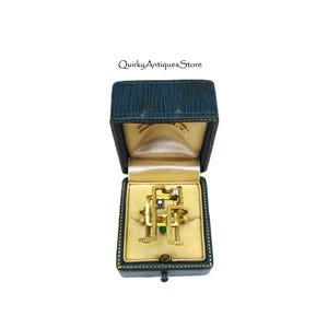 May include: A gold-toned ring with an abstract design, featuring small green and clear gemstones. The ring is displayed inside a vintage blue jewelry box with a gold satin interior. The box is open, showcasing the ring.