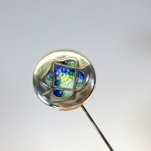 May include: Silver-toned hat pin with a round head featuring a colorful, abstract design. The design incorporates blue, green, and yellow elements within a geometric frame. The pin has a long, slender, dark metal shaft.