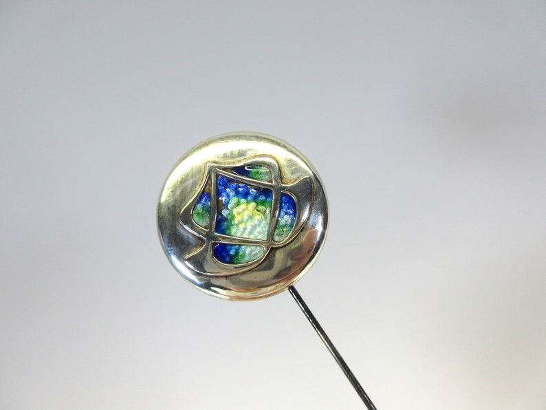 May include: Silver-toned hat pin with a round head featuring a colorful, abstract design. The design incorporates blue, green, and yellow elements within a geometric frame. The pin has a long, slender, dark metal shaft.