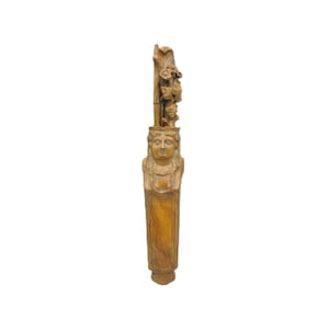 May include: A tall, light brown wooden carving featuring a central figure with a sculpted face and braided hair. Above the head, the wood is intricately detailed with carved floral elements and foliage, extending upward in a vertical, slender column.