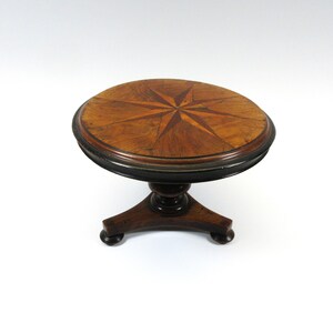 May include: A round wooden table with a starburst inlay in the top. The table has a dark brown finish and a pedestal base with three legs.