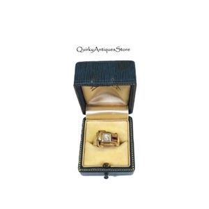 May include: A vintage gold ring with a square, multi-stone design, presented in a dark blue jewelry box with a gold interior. The ring features a central clear stone and two red stones. The box is open, showcasing the ring.