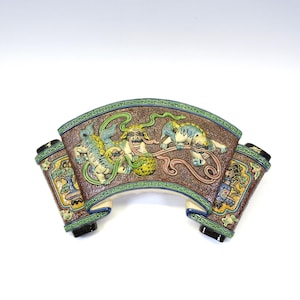 May include: A decorative ceramic scroll with a brown background and green trim. The scroll features a detailed relief of mythical creatures in shades of blue, green, and yellow. The ends of the scroll are rolled up, with black accents.