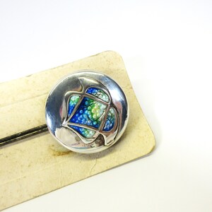 May include: A round, silver-toned brooch with an abstract design. The center features a mosaic of blue, green, and white beads. The brooch is set on a vintage, cream-colored card with a black pin.