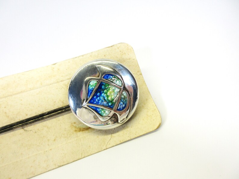 May include: A round, silver-toned brooch with an abstract design. The center features a mosaic of blue, green, and white beads. The brooch is set on a vintage, cream-colored card with a black pin.