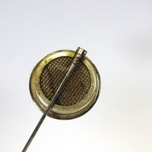May include: A vintage metal brooch with a circular design. The brooch features a mesh screen within a circular frame, with a long pin extending from the back. The frame has a worn, aged appearance.
