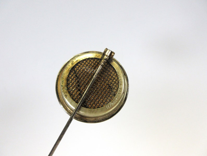 May include: A vintage metal brooch with a circular design. The brooch features a mesh screen within a circular frame, with a long pin extending from the back. The frame has a worn, aged appearance.