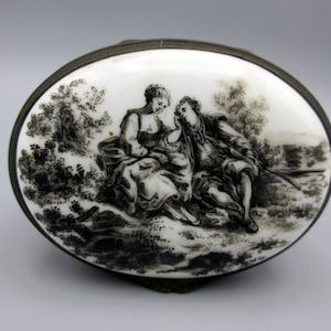 May include: An oval-shaped, silver-rimmed trinket box with a white porcelain lid. The lid features a black and white illustration of a couple sitting in a wooded area.
