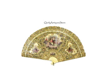 May include: A vintage hand fan with a gold and white paper design. The fan features a painted scene of a couple in a garden setting. The fan is closed and has a bone handle.