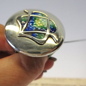 May include: A silver-toned brooch with a circular design. The center features a leaf-like shape with blue, green, and yellow enamel. The brooch is held by a hand, showcasing the intricate details and craftsmanship of the jewelry.