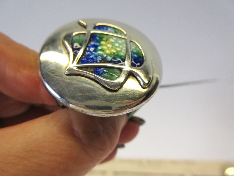 May include: A silver-toned brooch with a circular design. The center features a leaf-like shape with blue, green, and yellow enamel. The brooch is held by a hand, showcasing the intricate details and craftsmanship of the jewelry.