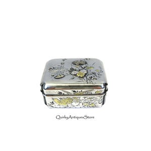 May include: A silver rectangular box with floral and bird designs. The box features gold accents on the flowers and a bird. The lid and sides are decorated with detailed engravings. The text "QuirkyAntiquesStore" is visible.