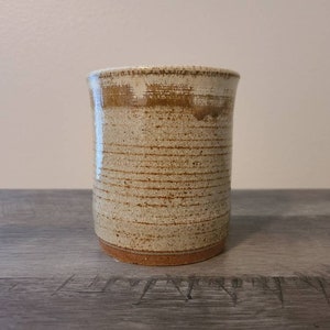 May include: A handmade ceramic mug with a cylindrical shape. The mug features a speckled beige exterior with horizontal brown lines and a brown band near the top. The base is a darker brown color. This rustic mug is perfect for coffee or tea.
