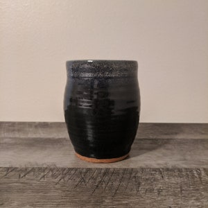 May include: A handmade, dark blue ceramic tumbler with a textured surface. The tumbler has a slightly flared shape and a natural, unglazed base. The rim has a speckled, lighter blue finish. The tumbler is sitting on a wooden surface.