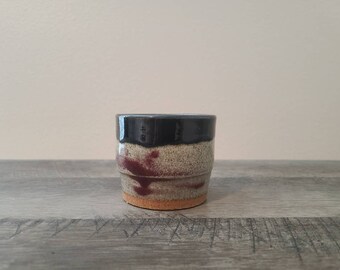 Könnte beinhalten: Eine kleine, handgefertigte Keramiktasse mit dunkelblauem Rand, beigefarbenem, gesprenkeltem Körper und weinroten Akzenten. Die Tasse hat eine strukturierte Oberfläche und eine natürliche, erdige Ästhetik. Der Boden ist hellbraun.