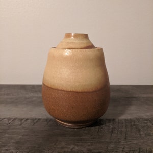 Op de afbeelding: Een handgemaakte keramische pot met een deksel. De pot heeft een afgeronde vorm met een lichtbruine bovenste helft en een donkerbruine onderste helft. Het deksel heeft een bijpassende bruine kleur. De pot staat op een houten oppervlak.
