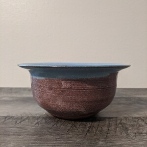 May include: A handmade ceramic bowl with a wide, flared rim. The upper portion is a light blue, transitioning to a textured, reddish-brown body. The bowl is designed for serving or display.