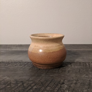 May include: A small, handcrafted ceramic pot with a rounded body and a flared rim. The pot features a two-tone color scheme, with a light tan upper section and a darker brown lower section. The pot is likely used for decorative purposes.