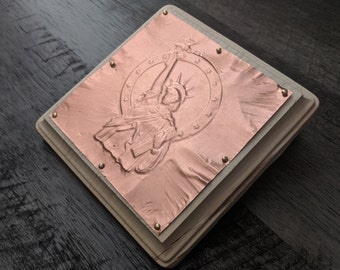 May include: A square, copper-colored metal plate with an embossed image of the Statue of Liberty, mounted on a wooden base. The plate is secured with small brass-colored rivets. The design features the statue holding a torch and a circle of stars.