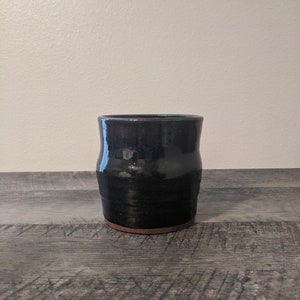 May include: A handmade black ceramic tumbler with a slightly flared rim and a textured, dark brown base. The glossy finish reflects light, highlighting the handcrafted nature of the pottery. The tumbler sits on a gray wooden surface.