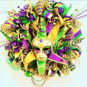 Puede incluir: Una corona de Mardi Gras con un esquema de color dorado, verde y morado. La corona presenta un sombrero de bufón y una máscara dorada y morada con una cara blanca.