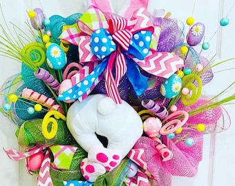May include: A colorful Easter wreath with a white bunny rabbit, pink bunny ears, a pink and blue bow, and various Easter decorations, including eggs, ribbons, and swirls.