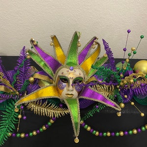 Puede incluir: Un centro de mesa con temática de Mardi Gras con una máscara de bufón púrpura, verde y dorada, helechos verdes y morados, cuentas doradas y moradas y adornos dorados y verdes.