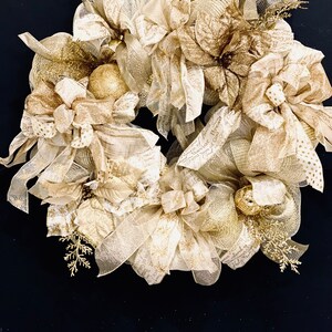 Puede incluir: Una corona de Navidad dorada y marfil con una variedad de cintas, lazos y adornos. La corona está decorada con una flor de pascua y tiene una cinta blanca colgando de la parte superior.
