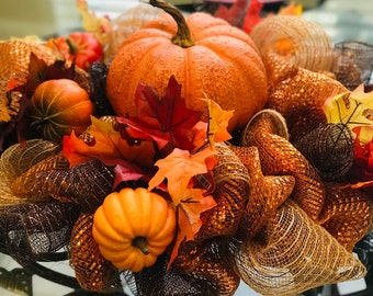 May include: A fall-themed centerpiece with a large orange pumpkin, smaller pumpkins, and brown and orange mesh ribbon. The centerpiece is decorated with fall leaves in shades of red, orange, and yellow.