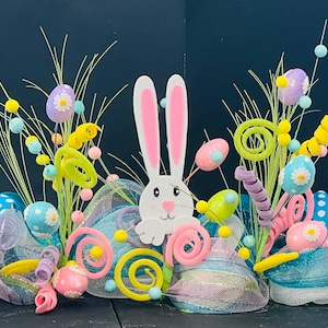 May include: A colorful Easter wreath with a white bunny face in the center. The wreath is decorated with pastel pink, blue, yellow, and green mesh, ribbons, and felt eggs.