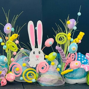 May include: A colorful Easter wreath with a white bunny, green grass, pink, blue, and yellow Easter eggs, and white and pink polka dot ribbon.