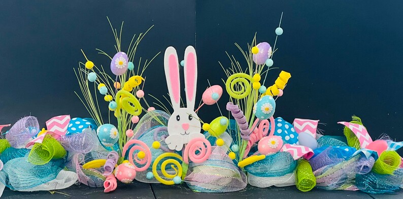 May include: A colorful Easter wreath with a white bunny, green grass, pink, blue, and yellow Easter eggs, and white and pink polka dot ribbon.