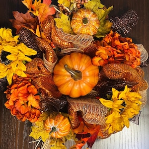 Puede incluir: Una corona de otoño con una gran calabaza naranja en el centro, rodeada de calabazas más pequeñas, girasoles y flores naranjas y amarillas. La corona está adornada con hojas de otoño y malla decorativa.