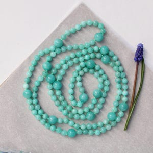 Puede incluir: Un collar largo de cuentas turquesas con cuentas redondas de diferentes tamaños. El collar está dispuesto en un patrón en espiral sobre una superficie gris claro. Una pequeña flor morada con un tallo verde se coloca a la derecha del collar.