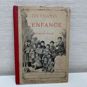 Op de afbeelding: Een vintage boek getiteld "Les Chants de L'Enfance" van Claude Auge. De kaft heeft een rode rug en een illustratie van kinderen die in een klaslokaal zingen.