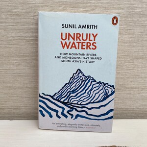 May include: A book cover with a blue and white illustration of a mountain range. The title of the book is "Unruly Waters: How Mountain Rivers and Monsoons Have Shaped South Asia's History" by Sunil Amrith.