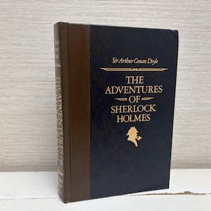 Könnte beinhalten: Ein dunkelblaues Hardcover-Buch mit goldenen Lettern. Der Titel lautet "The Adventures of Sherlock Holmes" von Sir Arthur Conan Doyle. Das Buch zeigt eine Silhouette von Sherlock Holmes mit einer Lupe.