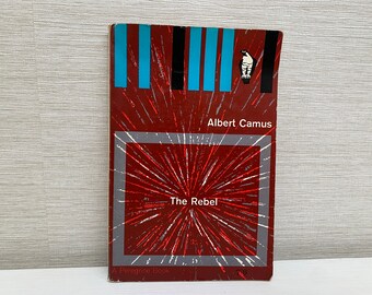 Könnte beinhalten: Ein Vintage-Taschenbuch mit dem Titel "The Rebel" von Albert Camus. Das Cover ist überwiegend rot mit einem weißen und grauen Burst-Design. Es zeigt blaue und schwarze vertikale Streifen oben und eine kleine Vogelillustration.